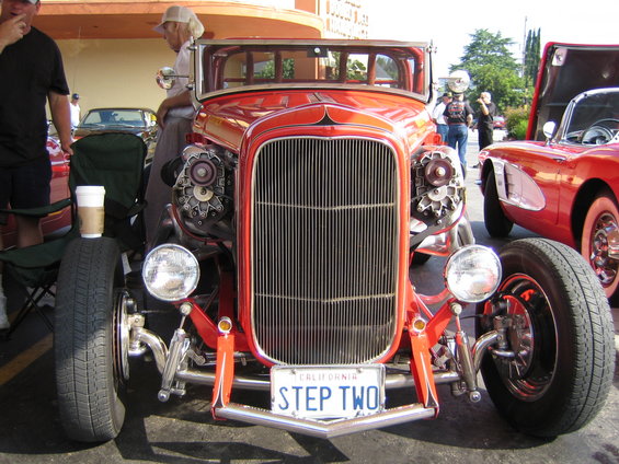 Front view of a red hot rod with a large chrome grille and dual engines visible on the sides. California license plate reads 'STEP TWO'. A red Corvette is parked to the right