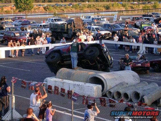 Off-road vehicle flipped on its side atop concrete pipes, with a crowd watching