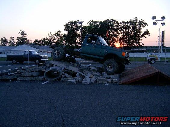 Green pickup truck attempting an obstacle course, with a black pickup truck parked nearby. Sunset in the background