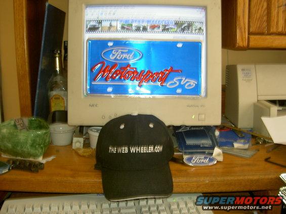Computer screen displaying a Ford Motorsport SVT logo and a row of small images at the top. A black baseball cap with 'THE WEB WHEELER.COM' is in front of the screen