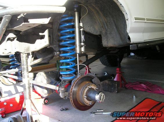 Front axle, suspension, and shock assembly on a vehicle lifted on jack stands. Blue coil springs are visible