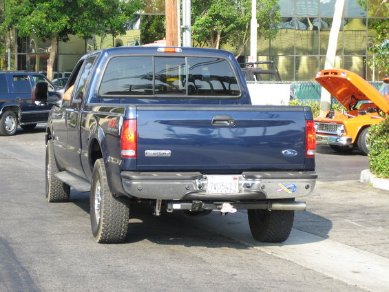 Blue Ford F-Series truck from behind, parked on street. Orange classic car with hood up in background