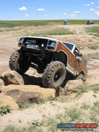 Brown and white SUV with large tires climbing over rocks on a dirt trail. Engine is exposed