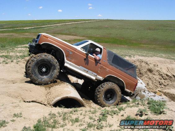 Brown Ford Bronco with big tires climbing over a concrete pipe in a dirt area
