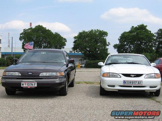 Dark sedan and white coupe parked side-by-side, front view. The sedan has a "W 1014 F" license plate