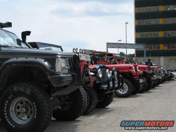Several Jeeps parked in a line, one black, others red. Building in background