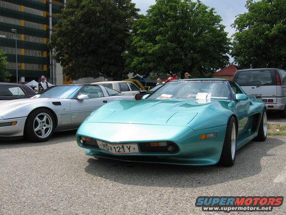Teal sports car with pop-up headlights and a license plate reading MO 121 Y. Silver sports car and a silver SUV are also visible