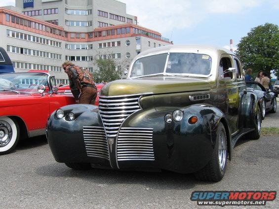 Front view of a heavily customized vintage car with a prominent chrome grille and rounded fenders, parked next to a red convertible