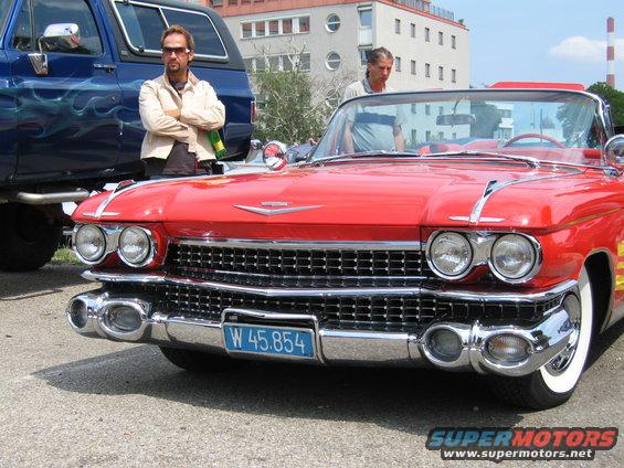 Red convertible car front view with chrome grille and dual headlights. Blue SUV in background