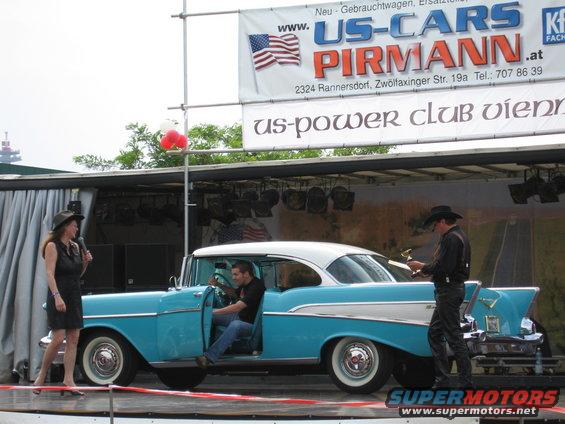 Blue and white 1957 Chevrolet Bel Air with driver's door open, woman with microphone standing nearby