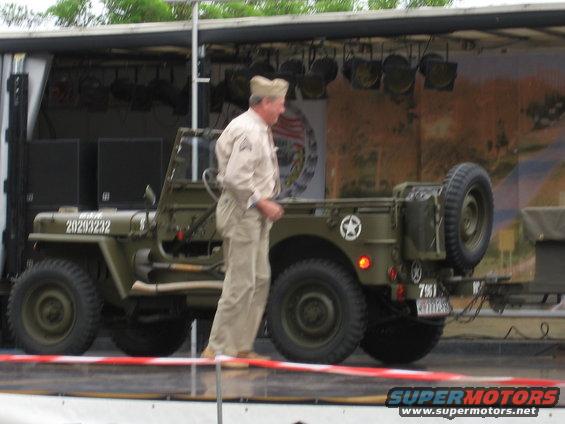 Green military jeep with a spare tire on the back and a man in a uniform standing next to it