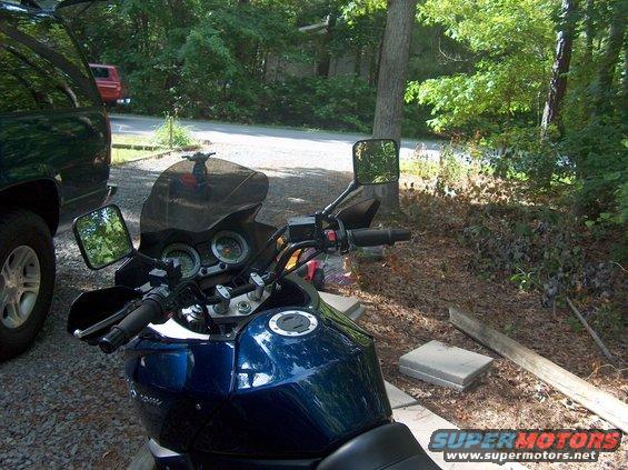 Blue motorcycle's handlebars, mirrors, and fuel tank are visible. A dark SUV is parked to the left