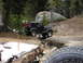 1989 Ford Bronco - A black Jeep Wrangler navigates a rough trail in a wooded area, with a person working on its underside.