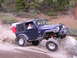 1989 Ford Bronco - A blue Jeep Wrangler navigates a rocky trail in a wooded area, equipped with extra fuel cans and storage.