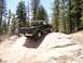 1989 Ford Bronco - A dark green pickup truck is shown driving on a challenging rocky terrain in a wooded area.