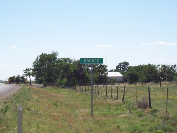 Green roadside sign reading BRONCO with a white building and trees in the background
