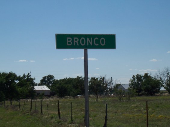 Green street sign that says BRONCO. Field and some buildings in background