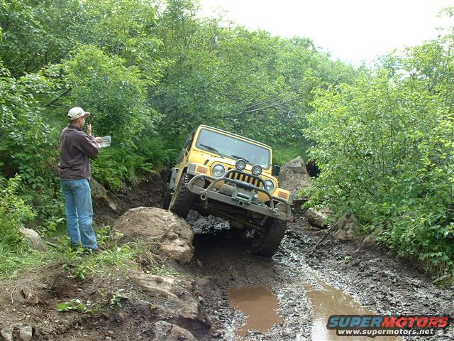 Yellow Jeep Wrangler navigating a muddy off-road trail with a person standing nearby holding a map