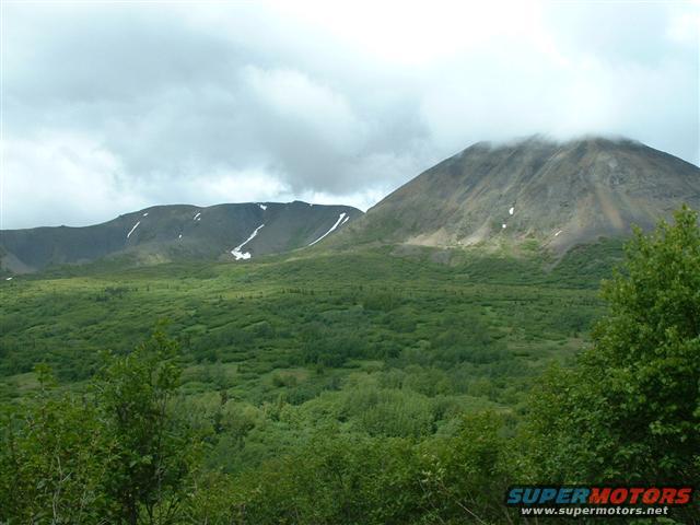 Mountainous landscape with green trees and cloudy sky, watermarked SUPERMOTORS.NET