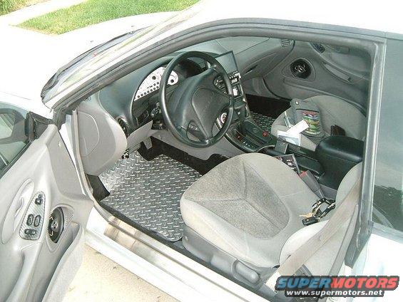 Car interior with diamond plate floor mats, grey seats, and a black steering wheel. A bag and tissues are on the passenger seat