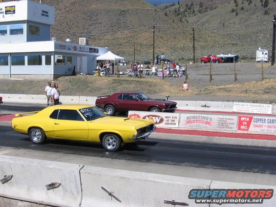 Yellow and maroon muscle cars racing on a drag strip with spectators in the background