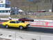 1972 Ford Mustang - Two classic muscle cars compete on a drag strip under a clear sky with mountains in the distance.