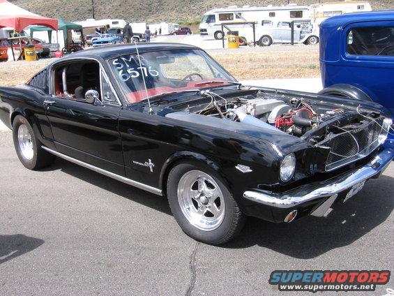 Black 1965 Ford Mustang fastback with red interior, hood open, showing engine, parked outdoors