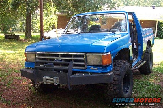 Blue pickup truck with aftermarket bumper and winch. It has large off-road tires and the driver's door is open