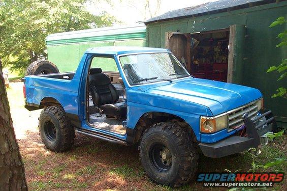 Blue Ford Bronco with aftermarket bumper and large tires, driver's door open, spare tire mounted in bed