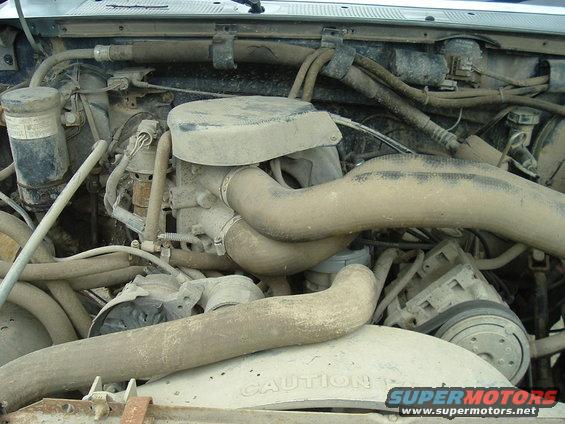 Dusty engine bay with hoses, air intake, and AC compressor visible. Text 'CAUTION' on fan shroud
