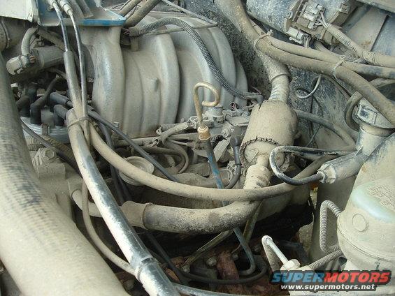 Engine bay with dusty intake manifold, spark plug wires, and hoses. Some text visible on a white container