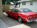 1974 Ford Maverick - A red 1974 Ford Maverick sedan with a black vinyl roof is parked in front of a white house.