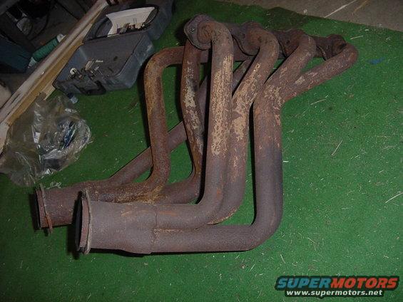 Rusty exhaust manifold with six pipes and two flanges, resting on a green surface