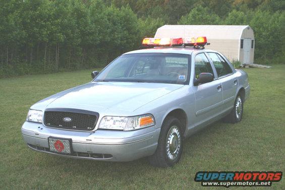 Silver 1998 Ford Crown Victoria police car with light bar parked on grass in front of a shed
