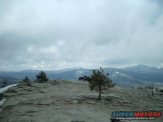 Two trucks parked on a rocky mountain overlook with trees and distant snow-capped mountains under a cloudy sky