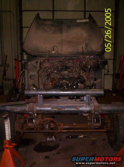 Jeep Wrangler chassis with engine removed, hood up, and front bumper modified. Vehicle is on jack stands in a garage
