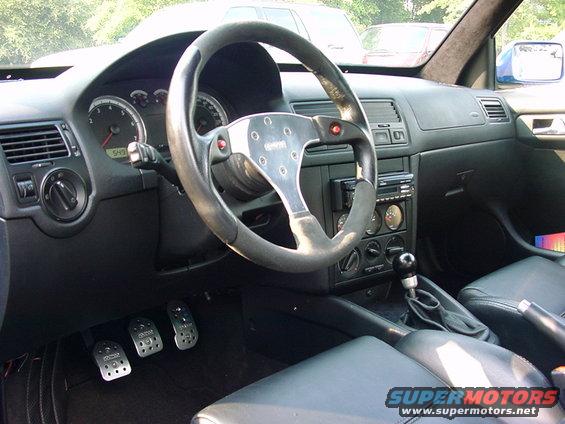 Interior view of a car, showing dashboard, aftermarket steering wheel, pedals, and gear shifter. A "SUPERMOTORS.NET" watermark is visible