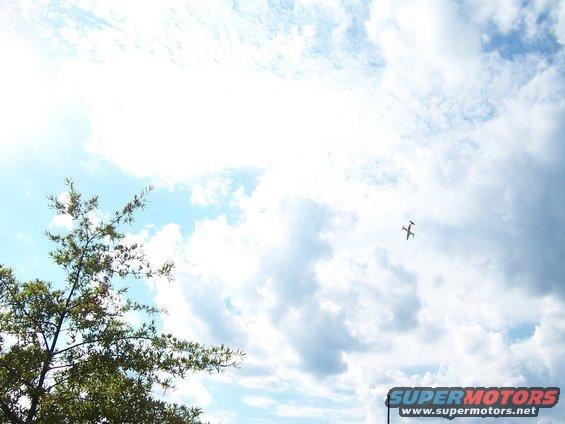 Small airplane flying in a cloudy sky, with tree branches in the foreground. Supermotors.net logo at bottom