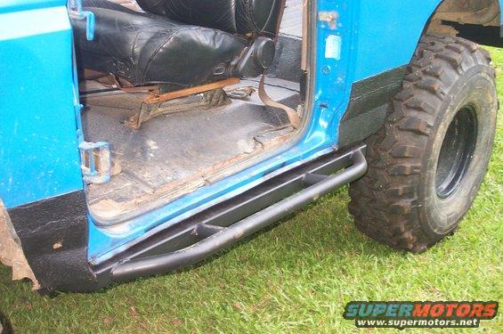 Close-up of aftermarket rock sliders installed on the side of a blue vehicle, showing the interior floor and a large off-road tire