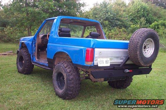 Blue 1987 Ford Bronco with aftermarket bumper and spare tire mounted on the back. It has large off-road tires and the doors are removed