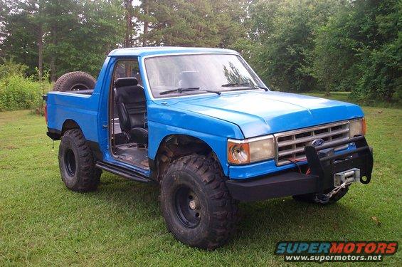 Blue 1987 Ford Bronco with lifted suspension and off-road tires. Driver's door is open, showing black interior. Winch mounted on front bumper
