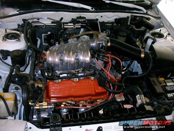 Engine bay with a V6 engine, chrome intake manifold, and red valve covers. Some hoses and wiring are visible