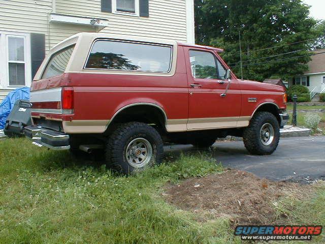 Red 1991 Ford Bronco, side view, tan top, lifted suspension, chrome bumper, parked on grass