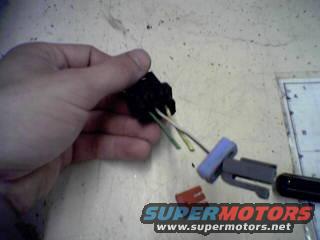 Hand holding a black automotive electrical connector with yellow, green, and white wires. A blue and gray connector piece is attached