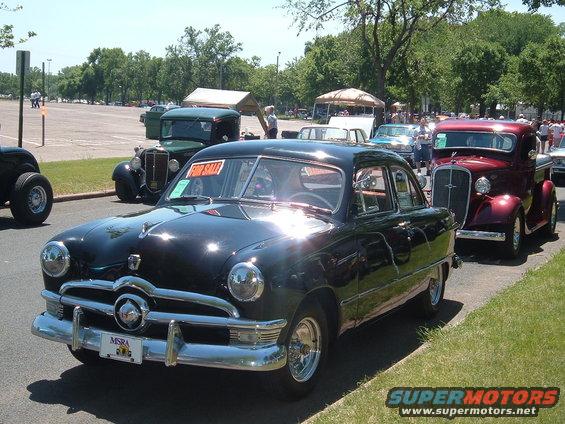 Black 1950 Ford sedan with a 'For Sale' sign in the window, parked on a street with other classic cars