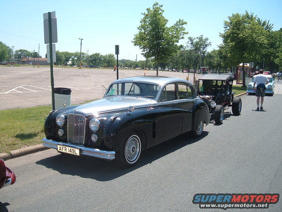 Black and silver sedan with a license plate reading AFR 149L, parked on asphalt next to a hot rod