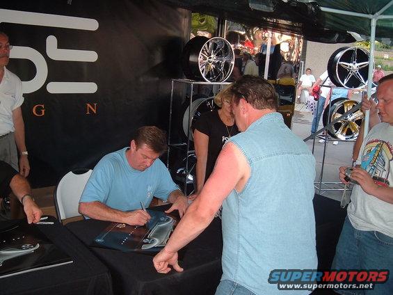 People signing autographs at a table with car wheels displayed