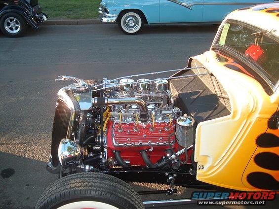 Hot rod engine bay with red V8 engine, chrome details, and flame paint on the body. A blue 1950s car is visible in the background