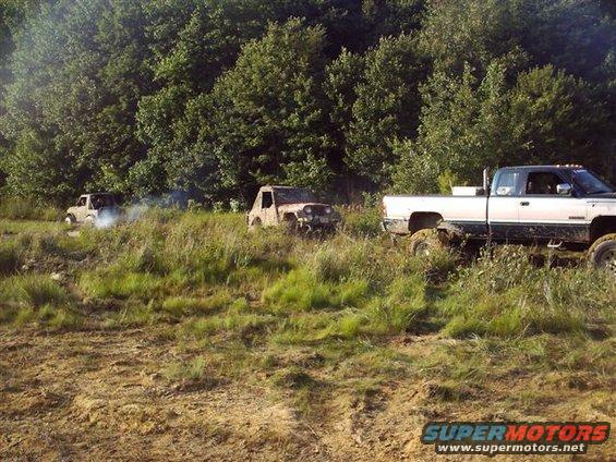 White and silver pickup truck with large tires in a muddy field, two other off-road vehicles in background