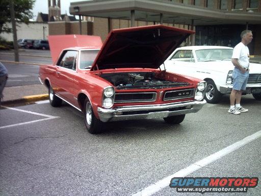 Red 1965 Pontiac GTO with hood open, parked next to a white car. A man stands between the cars